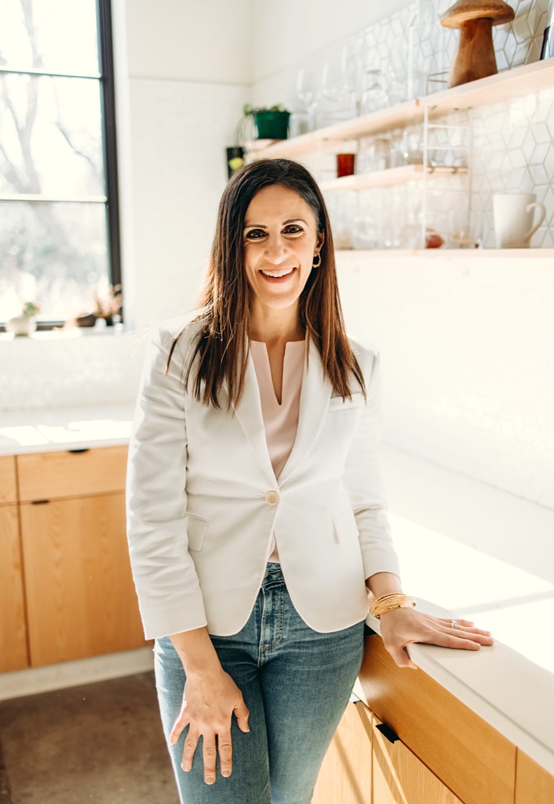 Nicole Runyon smiling in a white blazer, leaning on a sunlit kitchen counter